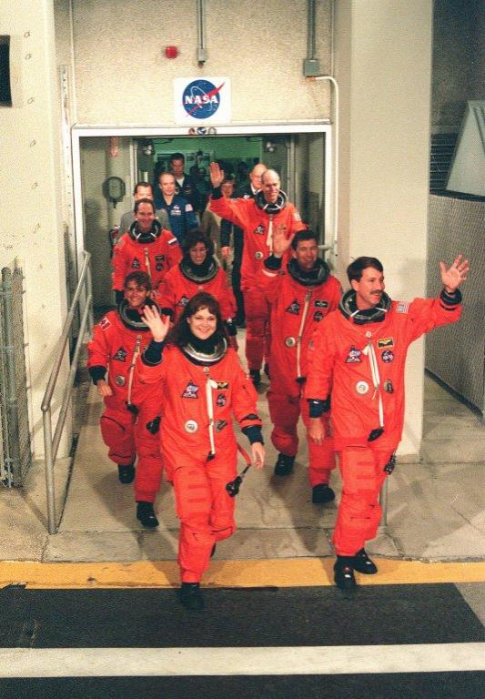 The STS-96 crew wave to onlookers as they walk out of the Operations and Checkout Building enroute to Launch Pad 39B and liftoff of Space Shuttle Discovery, targeted for 6:49 a.m. EDT. In their orange launch and entry suits, they are (clockwise from bottom left) Mission Specialists Tamara E. Jernigan, Julie Payette, Ellen Ochoa, Valery Ivanovich Tokarev and Daniel T. Barry, Pilot Rick D. Husband, and Commander Kent V. Rominger. Payette is with the Canadian Space Agency, and Tokarev is with the Russian Space Agency. STS-96 is a 10-day logistics and resupply mission for the International Space Station, carrying about 4,000 pounds of supplies, to be stored aboard the station for use by future crews, including laptop computers, cameras, tools, spare parts, and clothing. The mission also includes such payloads as a Russian crane, the Strela; a U.S.-built crane; the Spacehab Oceaneering Space System Box (SHOSS), a logistics items carrier; and STARSHINE, a student-involved experiment. It will include a space walk to attach the cranes to the outside of the ISS for use in future construction. Landing is expected at the SLF on June 6 about 1:58 a.m. EDT